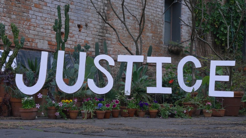 White JUSTICE letters among potted plants against a brick wall